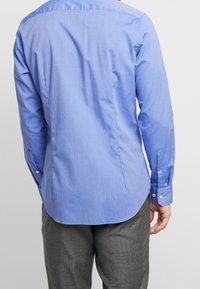 Modrá bavlněná košile s dlouhými rukávy, zadními sklady, zakřiveným lemem a zapínáním na knoflíky, v kombinaci se šedými vzorovanými kalhotami.