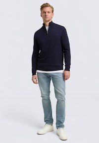 Granatowy sweter na zamek z długimi rękawami, wykonany z dzianiny. Połączony z jasnoniebieskimi jeansami i białymi trampkami, tworząc casualowy wygląd.