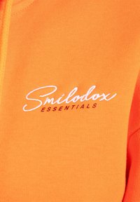 Hoodie orange avec un tissu texturé, une fermeture éclair à l'avant et le logo brodé "Smilodox ESSENTIALS" en blanc et rouge foncé.