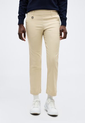 Persoon draagt beige rechte broek, witte sokken, witte sneakers en een marineblauwe top met lange mouwen, staand tegen een effen achtergrond.