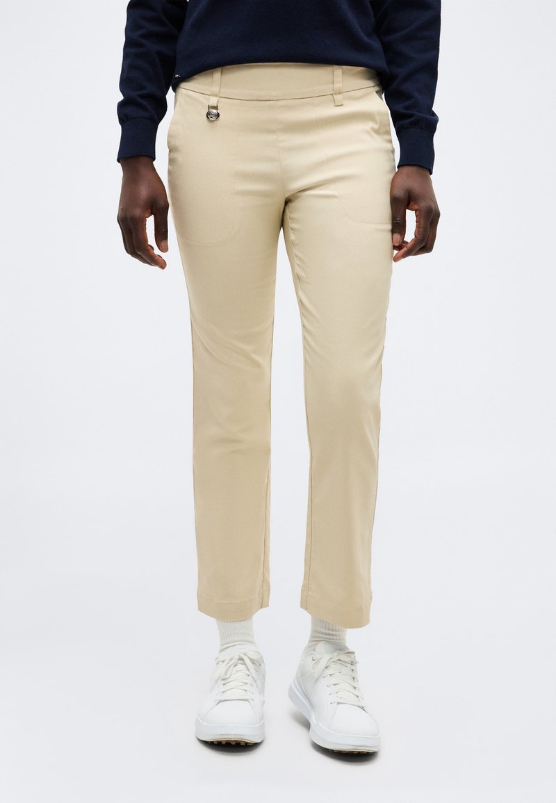 Άτομο που φοράει μπεζ παντελόνι με ίσια μπατζάκια, άσπρες κάλτσες, άσπρα αθλητικά παπούτσια και μπλε μακρυμάνικη μπλούζα, στέκεται μπροστά από ένα απλό φόντο.