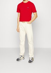 T-shirt rosso a maniche corte con un piccolo logo, abbinato a pantaloni color crema e sneakers mimetiche. Presenta una vestibilità standard e scollo rotondo.