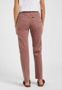 Persona vestida con jeans rectos color malva y una camisa blanca, mostrada desde atrás, de pie sobre un fondo claro liso.