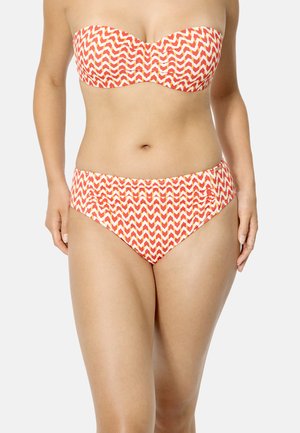 Mujer con top de bikini sin tirantes con patrón de zigzag en rojo, blanco y amarillo, y bragas de bikini de talle alto, sobre fondo liso.