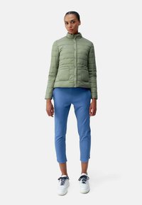 Groene gewatteerde jas met een hoge kraag en twee voorzakken, gecombineerd met blauwe tapered broek en witte sneakers met donkere veters.