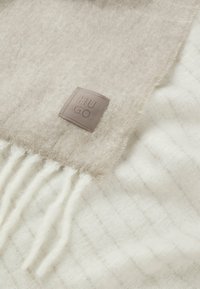 Beige uld tørklæde med en blød tekstur, som har et brunt læderlogo i patch og frynser langs kanten.