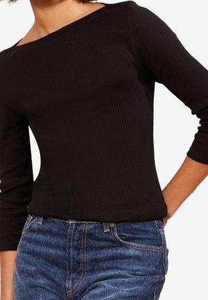 Femme portant un haut noir côtelé ajusté à manches longues, rentré dans un jean bleu taille haute, avec une main dans la poche.