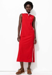 Robe sans manches rouge avec un col blanc, fentes latérales et accents blancs contrastants. Le mannequin porte des baskets marron.