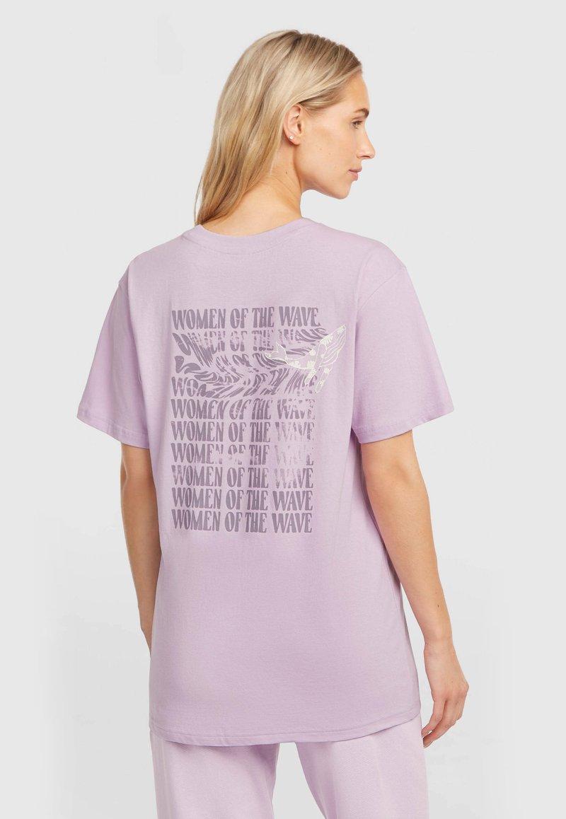 Μωβ βαμβακερό μπλουζάκι με γραφικό σχέδιο στην πλάτη που περιλαμβάνει επαναλαμβανόμενο κείμενο «ΓΥΝΑΙΚΕΣ ΤΟΥ ΚΥΜΑΤΟΣ» και μια απεικόνιση ενός πουλιού.