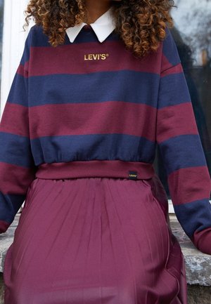 Osoba z kręconymi włosami ubrana w granatowo-bordową pasiastą bluzę Levi’s, biały kołnierzyk i pasującą plisowaną bordową spódnicę, siedzi na kamieniu.