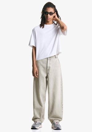 Weißes, übergroßes T-Shirt kombiniert mit hellgrauen, lockeren Jeans. Das Modell trägt schwarze Sonnenbrillen und graue Sneakers mit roten Akzenten.