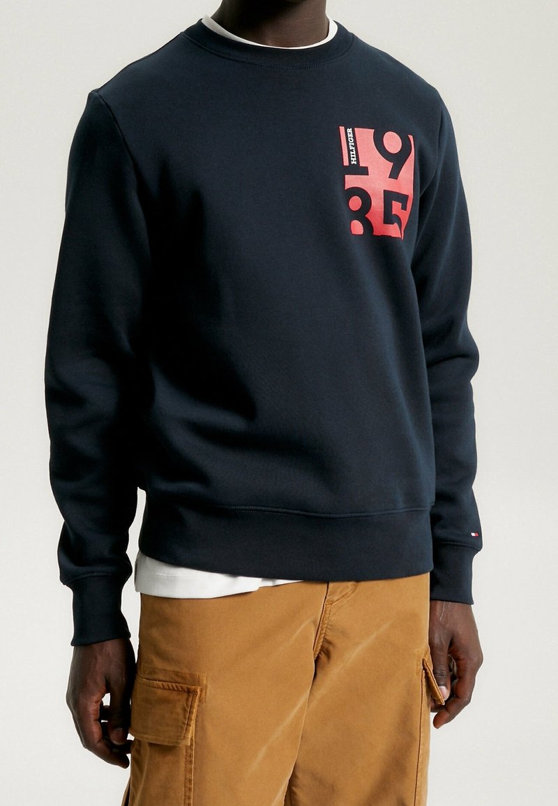 Hombre que lleva un sudadera azul marino con un gráfico rojo "1985" y pantalones cargo color beige, de pie contra un fondo claro y liso.