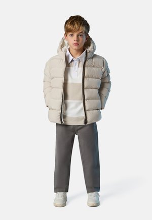 Doudoune beige clair avec capuche, coutures d'accent gris, polo blanc à rayures beiges, pantalon gris et baskets blanches.