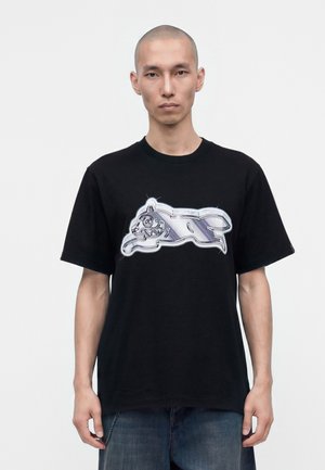 Mand med barberet hoved iført sort t-shirt med metalisk sølv løbende dyre grafik, stående mod en ensfarvet hvid baggrund.