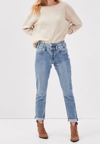 Nainen, joka on pukeutunut beigeen neuleeseen, joka on tupattu korkeavyötäröisiin vaaleansinihiin farkkuihin, joissa on käärityt lahkeet, ja ruskeisiin nubukista valmistettuihin nilkkasaappaisiin.