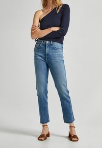 Žena v námořnickém topu na jedno rameno, s vysokým pasem modrými džíny s třepenými lemy a hnědými sandály na podpatku, stojí se zkříženýma rukama.