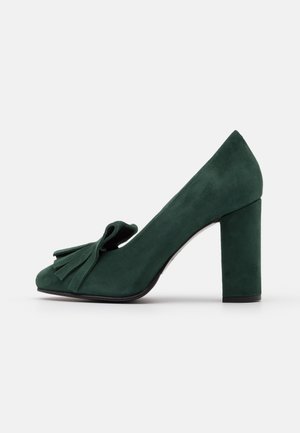 Chaussure à talon haut en daim vert avec un bout carré, un détail de nœud proéminent à l'avant et un talon épais pour la stabilité.