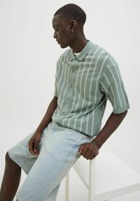 Lys grøn, stribet t-shirt med korte ærmer og knapper samt tekstureret stof, parret med lyseblå denim shorts. Model siddende på en hvid stol.