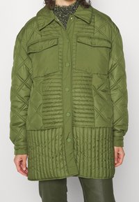 Chaqueta acolchada de color verde oliva con cierre de botones, dos bolsillos en el pecho y texturas variadas. Presenta patrones de costura en diamante y verticales.