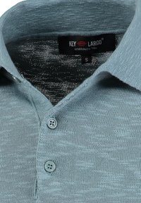 Lichtblauwe gebreide polo met een kraag, voorzien van twee knopen en een subtiele textuur. Merkkenlabel zichtbaar aan de binnenkant van de kraag. Maat: S.