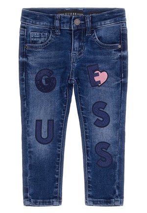 Jean pour enfants bleu foncé avec poches avant et lettres décoratives bleu marine formant le mot « GUESS » ainsi qu’un patch en forme de cœur rose sur la cuisse droite.