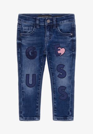Jean pour enfants bleu foncé avec poches avant et lettres décoratives bleu marine formant le mot « GUESS » ainsi qu’un patch en forme de cœur rose sur la cuisse droite.