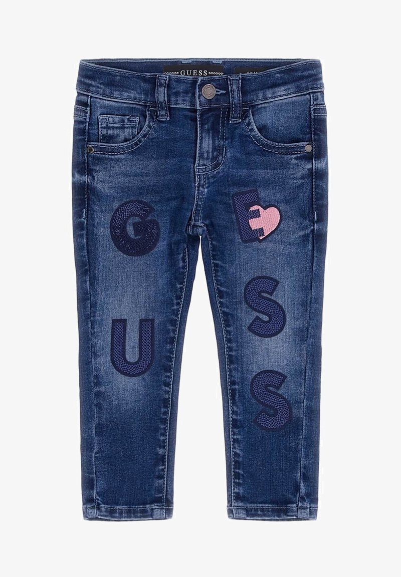 Jean pour enfants bleu foncé avec poches avant et lettres décoratives bleu marine formant le mot « GUESS » ainsi qu’un patch en forme de cœur rose sur la cuisse droite.
