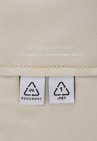 Beige fabric bag with a textured surface, featuring two labels indicating materials: “Doughnut” and “rPET.” Text states it’s made from recycled water bottles.