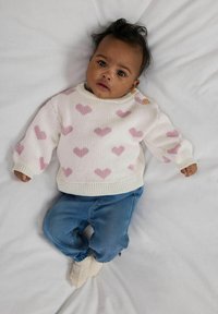 Sweter robiony na drutach w kolorze kremowym z różowymi wzorami serc, z długimi rękawami i detalem w postaci guzika. Połączony z niebieskimi dżinsami i kremowymi skarpetkami.
