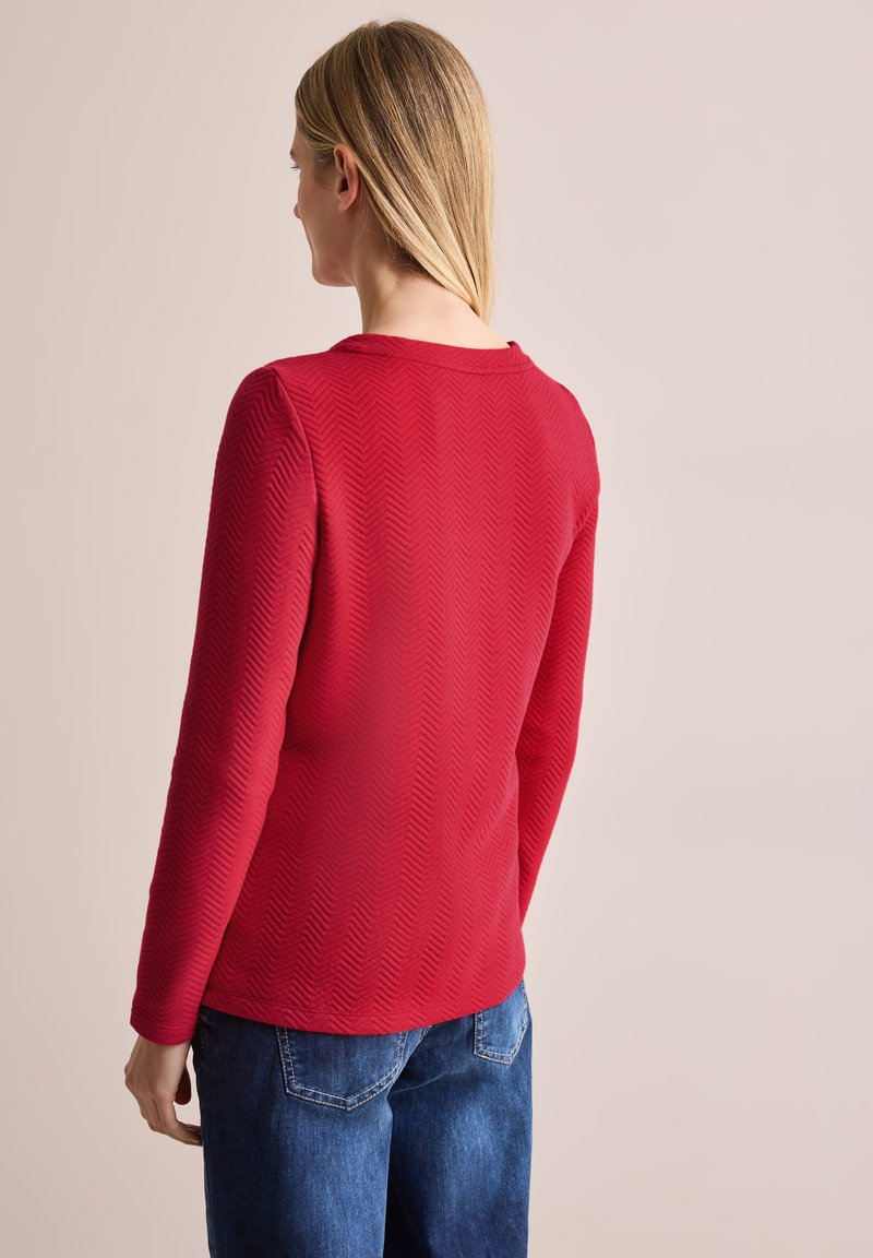 Rode langekoestop met een visgraatstructuur, ronde halslijn en een relaxte pasvorm, gecombineerd met blauwe jeans en een subtiele stikselafwerking.