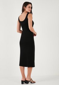 Musta midi-mekko, jossa on viehättävä siluetti, leveät olkaimet ja syvä selkäosa. Päällä mustat korkokenkä-sandaalit, joissa on kultaisia yksityiskohtia.