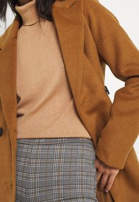 Manteau en mélange de laine couleur camel avec un col à revers, associé à un col roulé beige et une jupe grise à motifs avec un motif à carreaux.