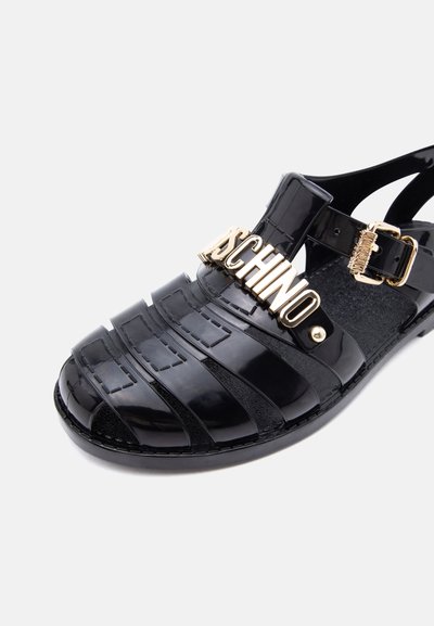 Sandale ouverte noire brillante avec boucle dorée et détail logo MOSCHINO doré sur la bride supérieure.