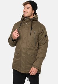Parka insulated de culoare verde măslin, cu glugă, închidere frontală cu fermoar și capse, două buzunare frontale și șnururi ajustabile. Căptușeală moale vizibilă.