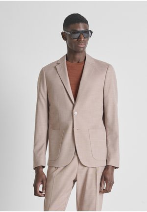 Mann in beigem Anzug und rostfarbenem Pullover, ergänzt durch große schwarze Sonnenbrillen, steht vor einem schlichten hellen Hintergrund.