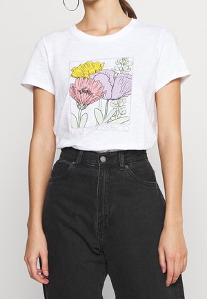 Vrouw draagt wit T-shirt met roze, gele en paarse bloemenprint, gecombineerd met hoge zwarte jeans en grote oorringen.