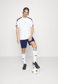 Vit atletisk t-shirt med svarta axelränder, marinblå shorts, vita strumpor med svarta detaljer och svarta fotbollsskor, stående bredvid en fotboll.