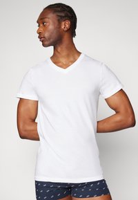 T-shirt en coton blanc à col V avec des manches courtes, design ajusté, texture lisse, présenté sur un modèle portant un short à motifs sombres, debout de profil.