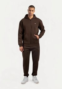 Sudadera marrón con cremallera y bolsillo frontal, combinada con pantalones jogger a juego. Ambas prendas están hechas de tela suave. El conjunto se completa con zapatillas blancas.
