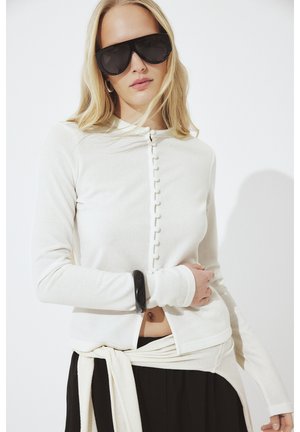 Žena nosi veľké čierne slnečné okuliare, biely zapínaný kardigán, čierny náramok a čierne nohavice so svetrom zviazaným v páse.