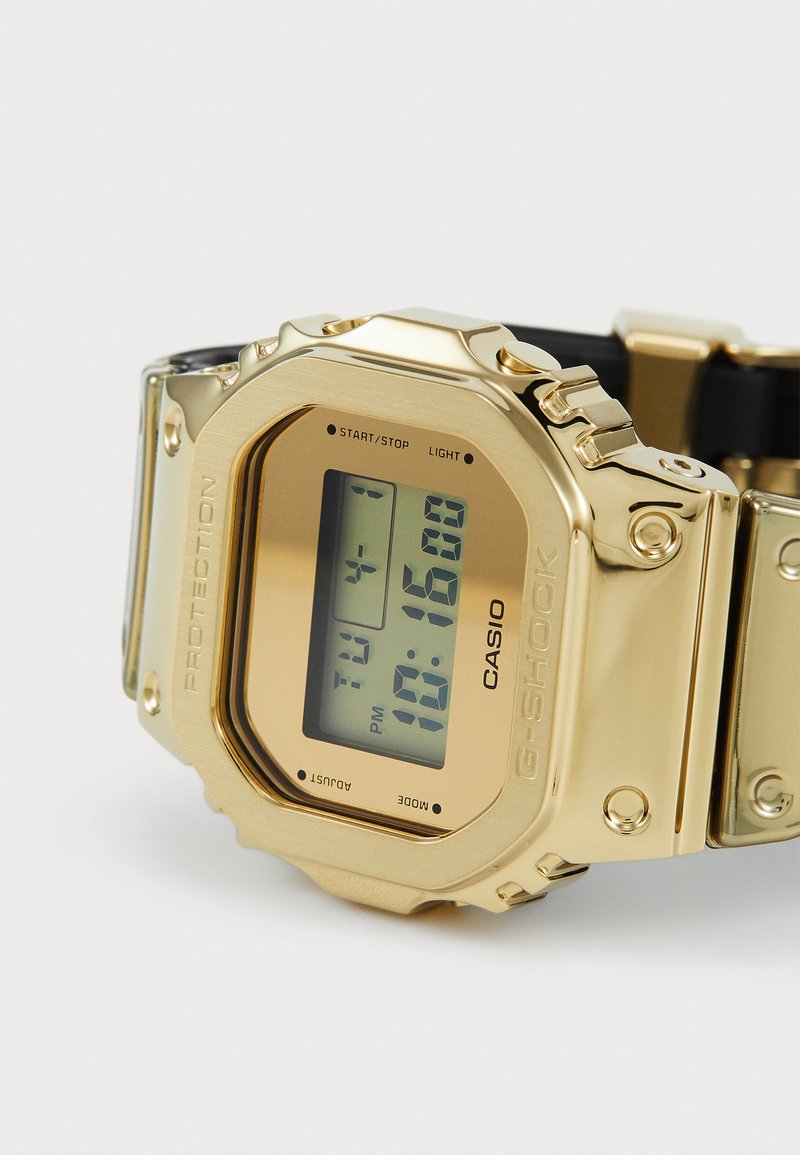 Orologio digitale con cassa in acciaio inossidabile dorato, cinturino in plastica nera e display LCD che mostra l'ora e le impostazioni della modalità.