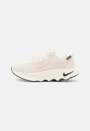 Lys beige Nike løbesko med mesh overdel, bølget tyk sål, sort Nike-logo på hælen og Gore-Tex mærkat nær kraven på ensfarvet baggrund.