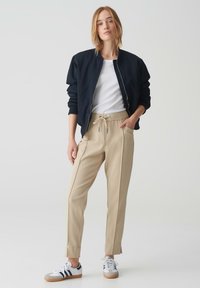Marine bomberjack, wit T-shirt, beige broek met trekkoord en witte sneakers met blauwe accenten en donkere strepen.