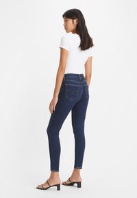 Dünne dunkelblaue Jeans mit Kontrastnähten, hoher Taille und Gesäßtaschen, kombiniert mit einem enganliegenden weißen T-Shirt und schwarzen Blockabsatzsandalen.
