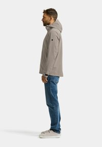 Hellgraue wasserdichte Jacke mit Kapuze, Seitentaschen und strukturiertem Stoff, getragen über blauen Jeans und grauen Turnschuhen.