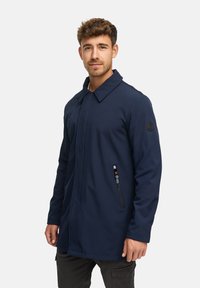 Marineblauer langen Mantel aus einem weichen, strukturierten Stoff; verfügt über einen Reverskragen, eine seitliche Zippertasche und ein Patch am linken Ärmel.
