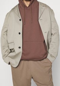 Beige leichte Jacke über einem braunen Kapuzenpullover mit Kordelzug, kombiniert mit braunen Hosen; verfügt über schwarze Knöpfe und dezente Nähdetails.