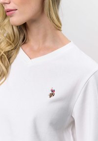 Biała bluza z dekoltem w serek wykonana z bawełny o gładkiej fakturze. Posiada haftowane kolorowe logo po lewej stronie klatki piersiowej.