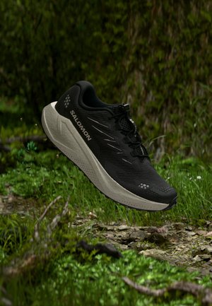 Scarpa da corsa Salomon nera in aria sopra erba rocciosa con sfondo di fogliame verde, mettendo in risalto suola e marchio laterale.