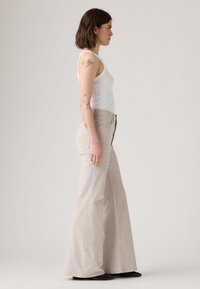 Jeans flare bege com uma cintura alta, costuras visíveis e dois bolsos traseiros. Combinados com uma t-shirt justa branca e sapatos pretos.